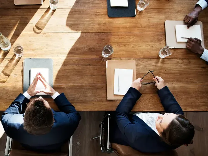 Overhead view of business professionals seated around a wooden conference table with notes, folders, and water glasses
